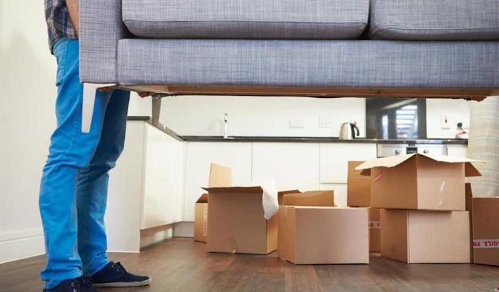 A man in jeans is picking up a grey couch; moving boxes are in the background