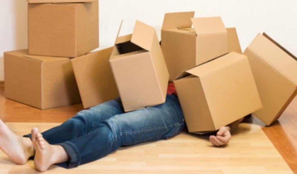 A person lies on the ground, completely covered by a stack of cardboard moving boxes