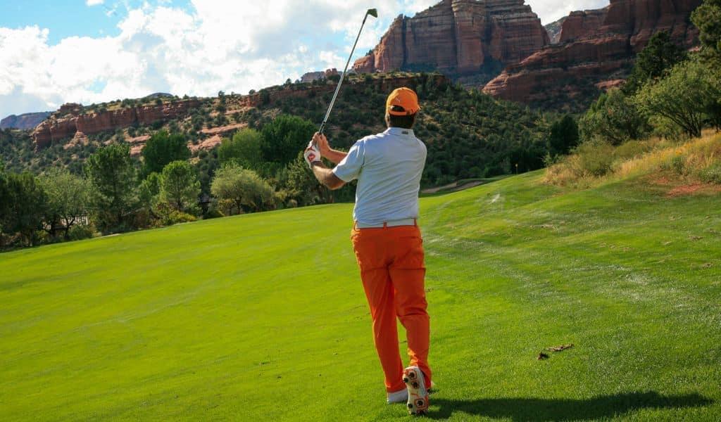 Man in red pans and white shirt playing golf on a green luscious course with mountains in the background