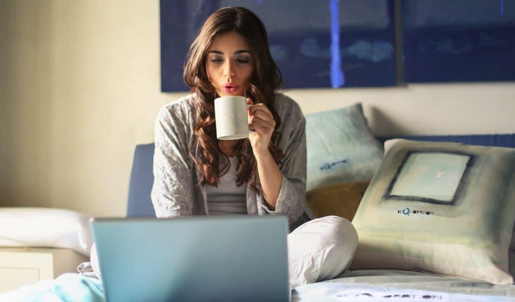 Woman wearing grey jacket sitting on bed with laptop and cup of coffee