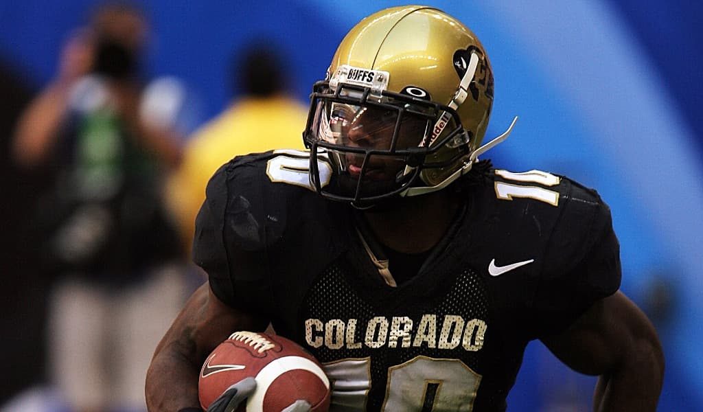 Colorado football player wearing black jersey and golden helmet while carrying a football on the field