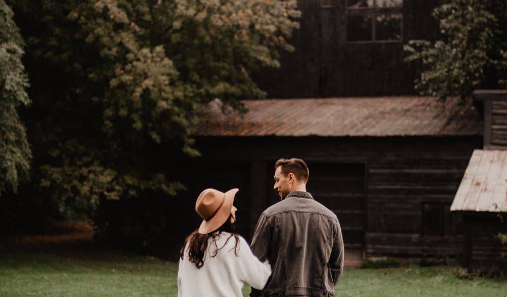 Woman in white jersey and hat standing with man in brown jacket on grass admiring a country home