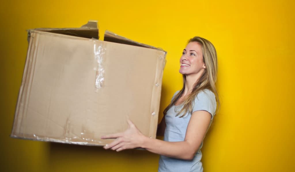 Woman wearing a grey shirt holding a brown cardboard box against a yellow background