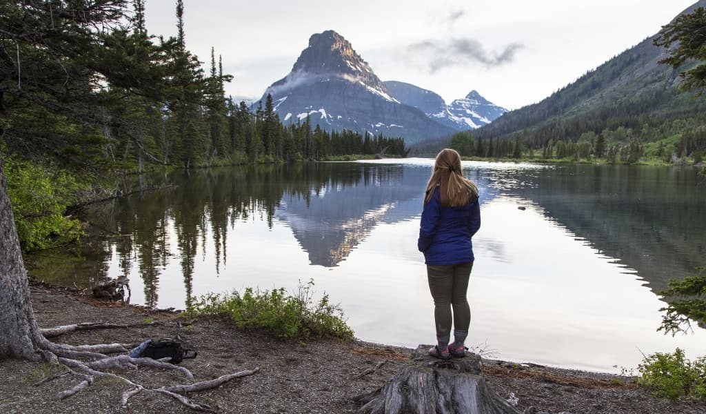 Woman in blue jacket overlooking a lake surrounded by trees and a mountain in the distance