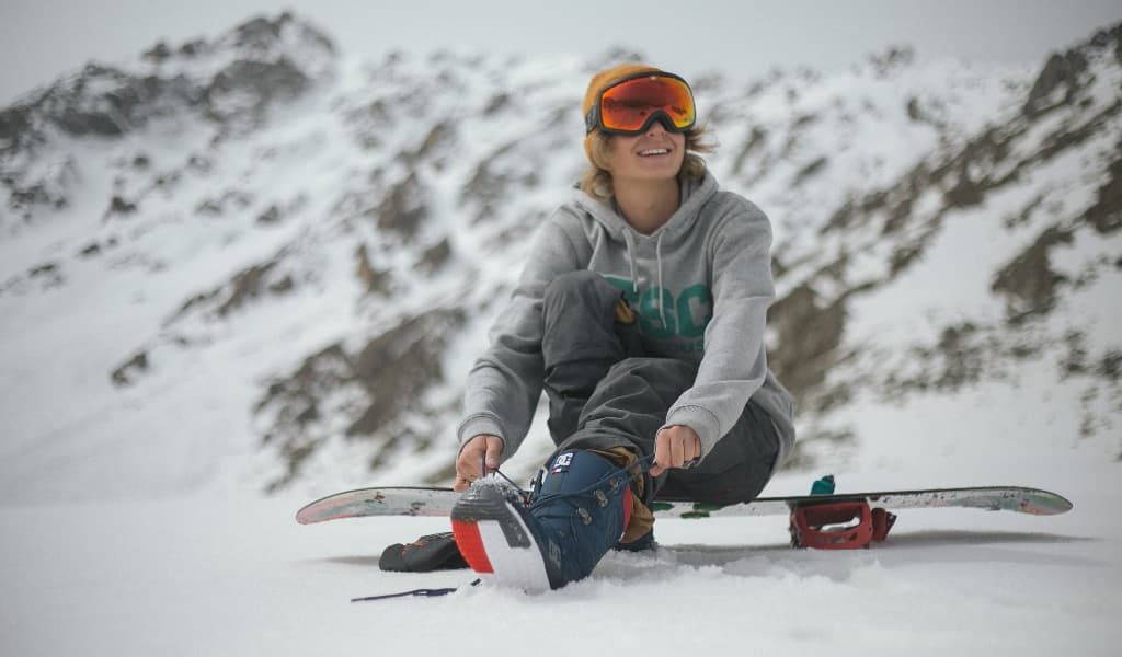 Woman wearing a grey hoodie sitting on a snowboard surrounded by snow