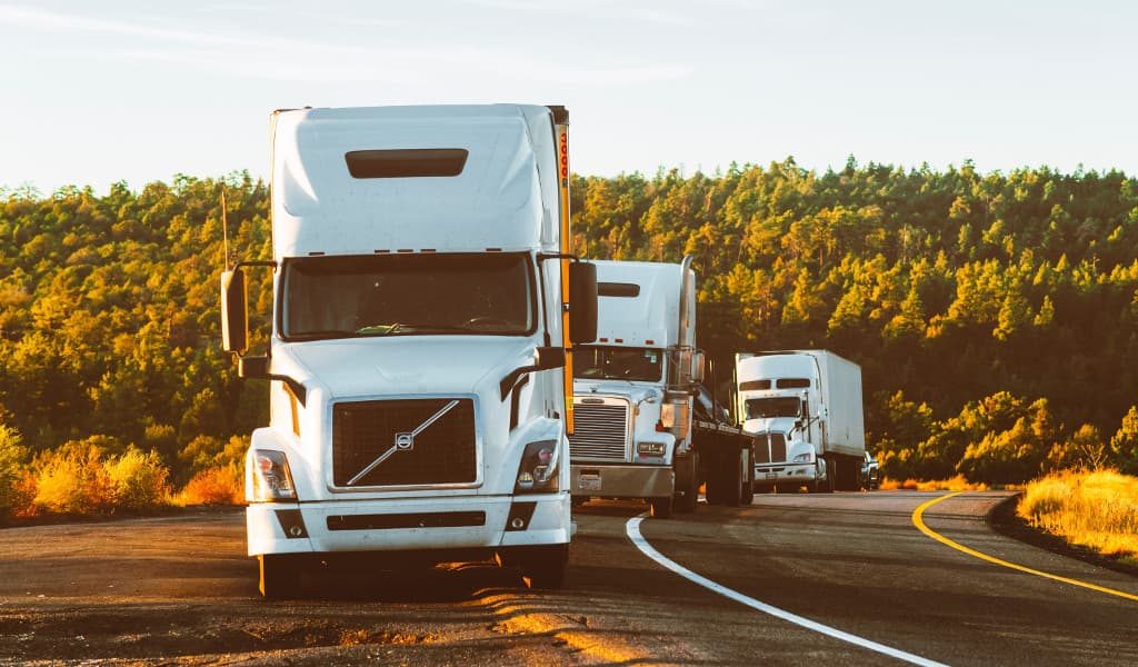 Three white moving trucks driving in succession on a road with trees in the background