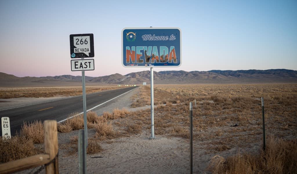 Blue ‘Welcome to Nevada Sign’ beside the main highway with mountains in the background